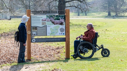 the photo shows two people, one of which is in a wheelchair, viewing a interpretation panel in the deer park